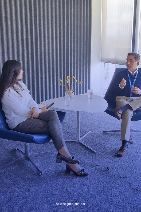 Mujer y Hombre con vestidos formales, teniendo una conversación en una oficina. Mesa de centro en vidrio con flores amarillas. Mujer entrevistando